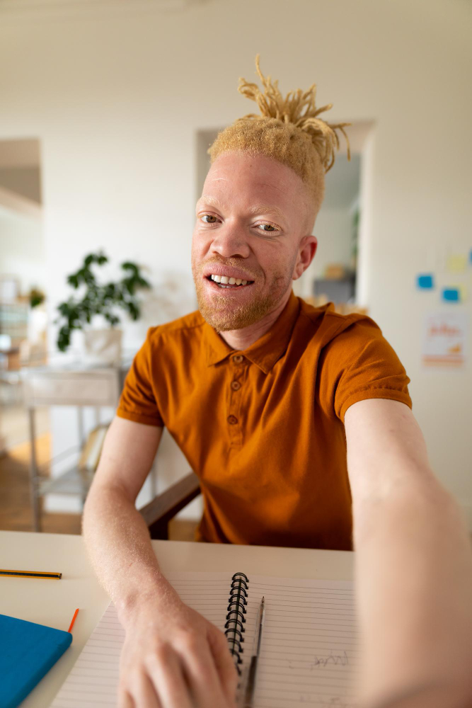 Man with albinism and blonde locs smiling at the camera while seated at a desk with an open notebook and pencils in front of him