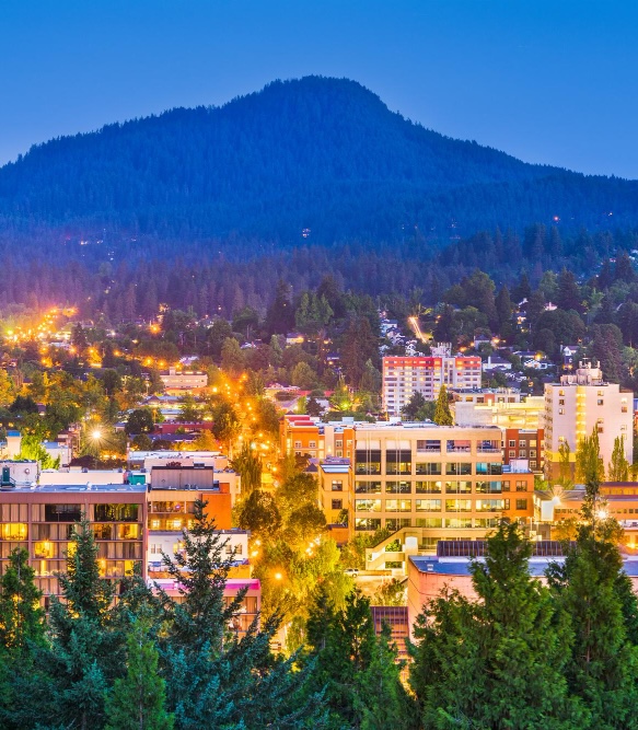 Twilight aerial view of Eugene, Oregon with city lights glowing warm against a darkening sky and a forested mountain rising in the background