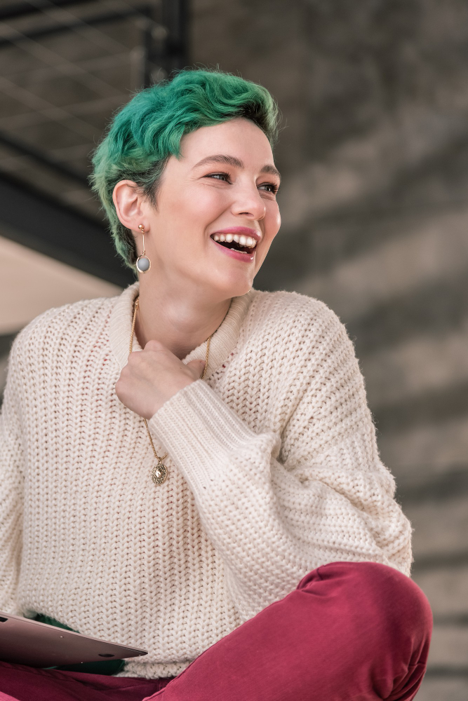 Woman with short green hair laughing openly with a laptop in her lap, wearing a cream knit sweater