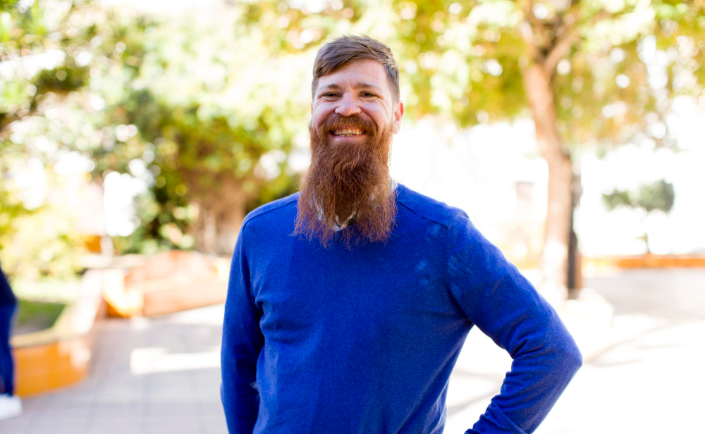 Smiling man with a full red beard wearing a bright blue sweater, standing in a sunny outdoor space with trees and planters behind him