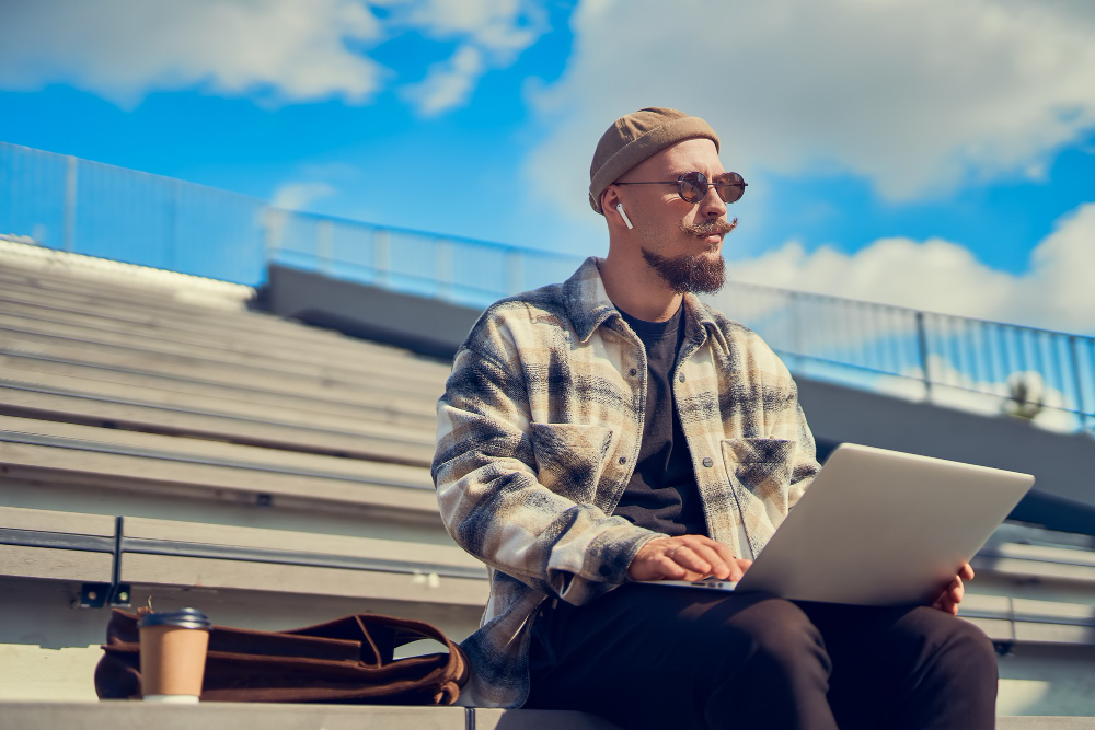 Person in a plaid jacket and sunglasses sitting on outdoor steps working on a laptop with a coffee cup and bag beside them