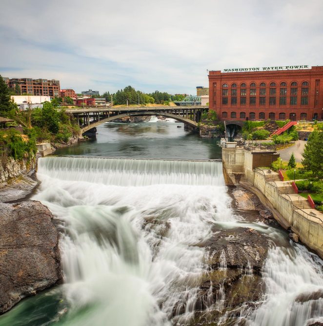 Spokane Falls rushing over rocky riverbanks in downtown Spokane, with the historic Washington Water Power building and a bridge in the background