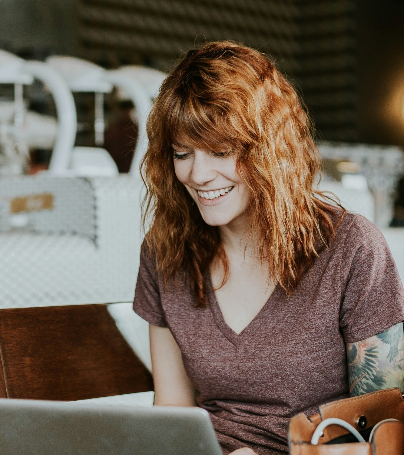 Woman with tattoos and curly red hair smiling while working on a laptop at a coffee shop, representing adults who seek virtual therapy in Austin